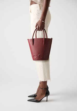 Woman holding burgundy leather tote bag with embossed logo, wearing cream cropped pants, black pointed-toe heels, and a beaded bracelet.