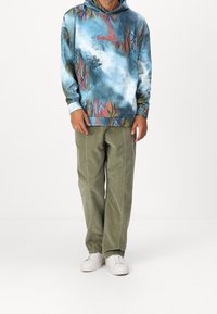 Uomo che indossa un hoodie multicolore blu con il testo rosso "Cole Colossi", pantaloni verde oliva e sneakers bianche, in piedi davanti a uno sfondo bianco.