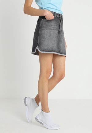 Personne portant une mini-jupe en jean noire délavée avec un ourlet arrondi, un haut bleu clair et des baskets blanches avec chaussettes, debout avec une main dans la poche.