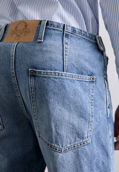 Jeans de culoare albastru deschis, cu croială dreaptă, prezentând o eticheta din piele pe spate și un singur buzunar posterior cu cusături vizibile.