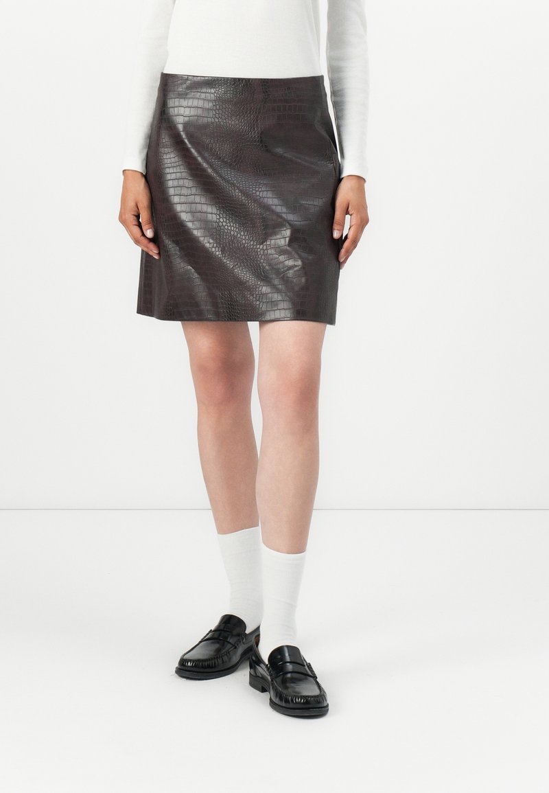 Femme portant une mini-jupe marron foncé en cuir synthétique imitation crocodile, un haut blanc à manches longues, des chaussettes blanches et des mocassins noirs, debout sur un fond blanc.