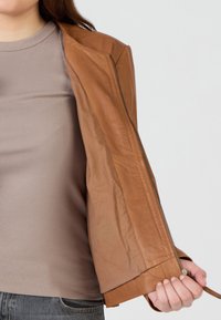 Hellbraune Lederjacke mit schmaler, taillierter Passform und Reißverschluss, partially geöffnet, um ein graues geripptes Oberteil darunter zu zeigen.