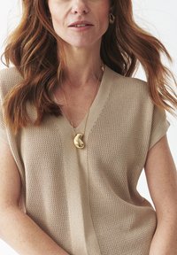 Top de punto beige con escote en V, mangas cortas y un patrón texturizado. Presenta un collar con colgante dorado y pendientes de aro.