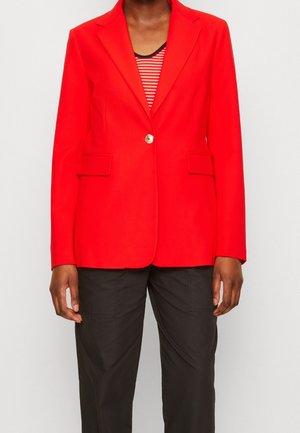 Rode blazer met een enkele gouden knoop, twee voorzakken, getailleerde pasvorm, gladde stof, gedragen over een zwart-wit gestreept shirt, gecombineerd met zwarte broek.