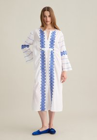 Abito bianco con ricami a diamante blu lungo il davanti e sulle maniche; presenta un collo a V e una silhouette rilassata. Scarpe flat blu.
