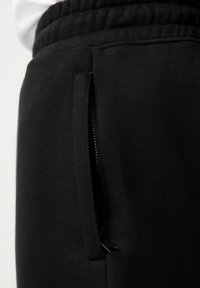 Crne trenirke imaju bočni džep s patentnim zatvaračem i elastičnu waistband. Tkanina je glatka i teksturirana, osmišljena za udobnost i fleksibilnost.