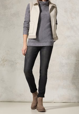 Femme portant un col roulé en maille grise, un gilet sans manches crème, un jean skinny noir et des bottines beiges, debout contre un mur neutre.