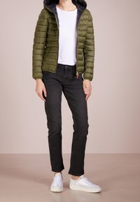 Olivengroene donzen jas met een capuchon, wit T-shirt, zwarte spijkerbroek en witte sneakers. De jas heeft een rits aan de voorkant en geribbelde manchetten.