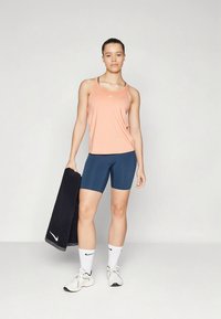 Egy nő barack színű sportmelltartóban és sötétkék biciklis shortban áll, egy fekete Nike törölközőt tartva, fehér Nike zoknikat és sportcipőt viselve, egy sima háttér előtt.