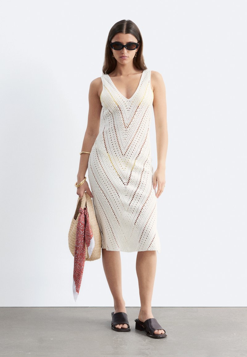 Femme portant une robe blanche en crochet sans manches avec un col en V, des lunettes de soleil noires, des sandales noires, des bracelets dorés et tenant un sac tissé avec un foulard rouge.