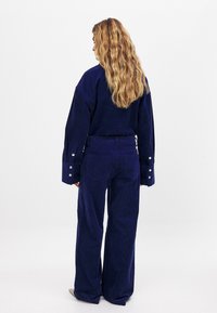 Jūrinės spalvos štrichinis komplektas, susidedantis iš per didelio marškinio su plačiais rankogaliais ir mygtukų akcentais, derinamas su atitinkančiomis plačiomis kelnėmis. Matomos audinio tekstūros detalės.