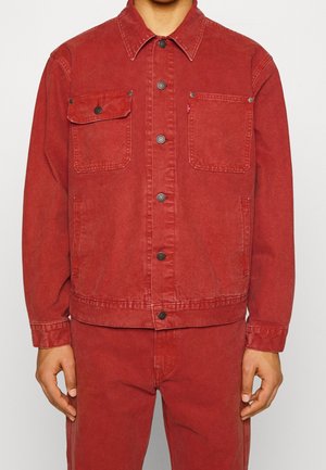 Mand iført matchende rød denimjakke med knaplukning på brystlommerne og røde denimbukser, stående foran en ensfarvet baggrund.