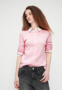 Ανοιχτό ροζ πλεκτό πουλόβερ με υφασμένο μοτίβο, πουκαμισοειδές με ρολά μανίκια πάνω από ένα λευκό πουκάμισο με γιακά, συνδυασμένο με σκούρο γκρι τζιν.