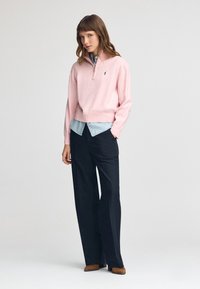Ljusrosa zip-up tröja i mjukt material, lager på lager över en blå chambrayskjorta, kombinerad med svarta vida byxor och bruna skor.
