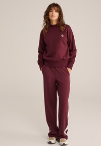 Egy nő áll, kezét a zsebében tartva, bordó pulóverben és hozzá illő nadrágban, fehér oldalcsíkokkal, bézs tornacipővel.