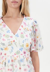 Robe florale blanche avec un décolleté en V, des manches courtes en dentelle transparente et une coupe fluide ornée de motifs floraux colorés dans des tons de rose, bleu et jaune.