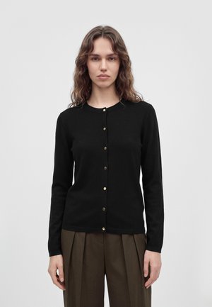 Un mannequin se tient devant un fond clair uni, portant un cardigan noir boutonné sur le devant et un pantalon marron.