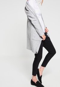 Grå ullkofta med öppen front, två framfickor, bärs över en vit skjorta och svarta byxor, tillsammans med svarta slip-on skor.