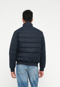Bărbat întors, purtând o jachetă din puf cu model matlasat, de culoare bleumarin, și blugi de culoare albastru deschis, pe un fundal alb simplu.
