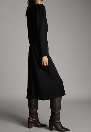 Femme portant une longue robe noire à manches longues et des bottes en cuir marron à talons jusqu'au milieu du mollet, debout devant un fond uni.