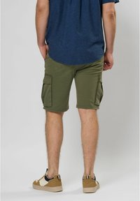 Olijfgroene cargoshorts met twee zijzakken, een rechte pasvorm en omgeslagen zoom, gecombineerd met beige sneakers en een blauwe shirt.