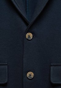 La жагавары синим текстурированным жакетом с вырезом, двумя передними карманами и характерными круглыми коричневыми пуговицами. Близкий план подчеркивает ткань и детали.