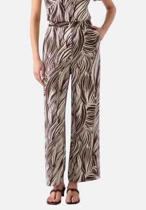 Donna che indossa pantaloni a gamba larga a vita alta con motivo astratto di foglie marroni e beige, abbinati a sandali con tacco marrone scuro.