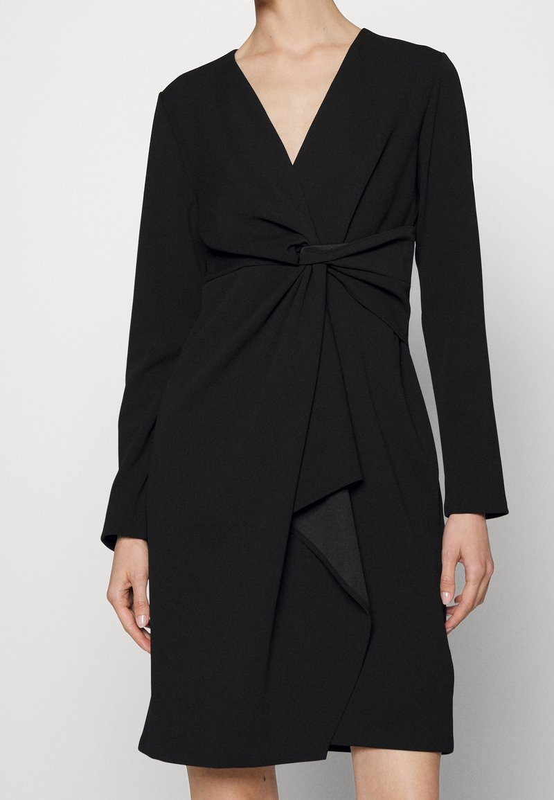 Robe portefeuille noire à manches longues, décolleté en V profond, détail torsadé à la taille, et ourlet avant superposé. Tissu lisse avec une silhouette structurée.