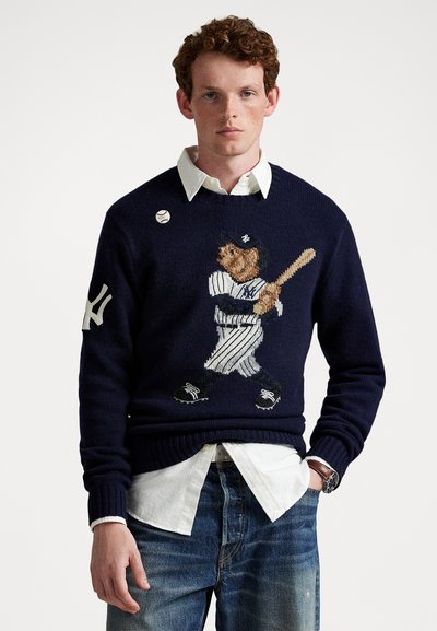 Mand iført navy sweater med broderet bjørn i baseballuniform, der holder bat, hvid skjorte med krave, blå jeans og ur.