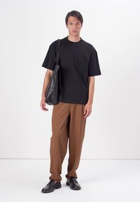 T-shirt noir à manches courtes avec une poche poitrine, associé à un pantalon ample marron et des mocassins noirs. Le modèle porte un sac à bandoulière noir.
