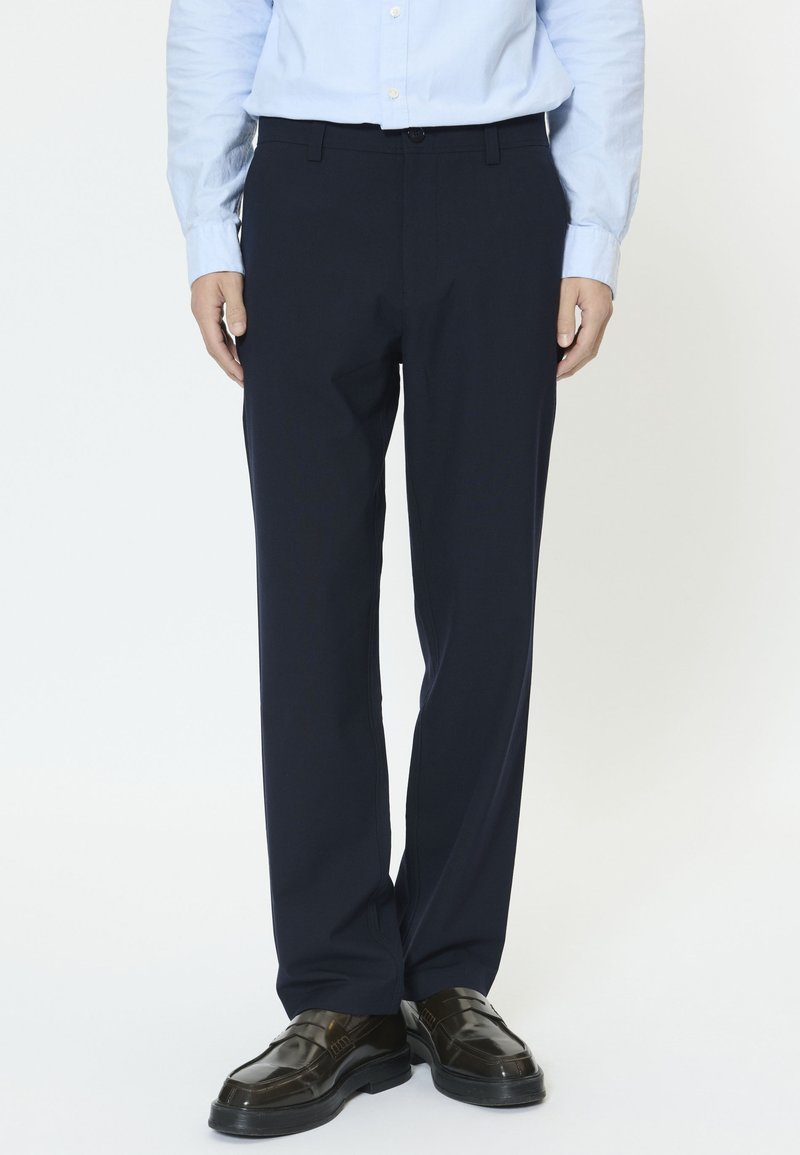 Pantalon bleu marine à coupe droite, fabriqué en tissu lisse, avec un devant plat, des passants pour ceinture et des poches latérales. Porté avec des chaussures noires brillantes.