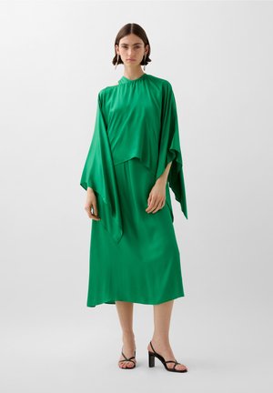 Vestido de satén verde con un cuello alto, mangas amplias y una silueta fluida, combinado con sandalias de tacón alto negras con tiras.