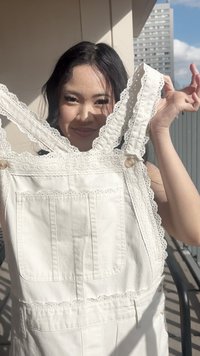 Jeune femme tenant une salopette blanche ornée de dentelle au soleil sur un balcon avec des bâtiments en arrière-plan.