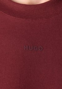 Gros plan sur un tissu côtelé couleur bordeaux avec le logo "HUGO" en relief centré sous l'encolure d'un vêtement à col rond.