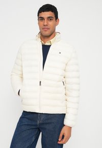 Valge paksust materjalist jakk horisontaalse tikandiga, ees tõmblukuga, küljetaskute ja väikese logoga rinnal. Kantud tumesinise kampsuni peal.