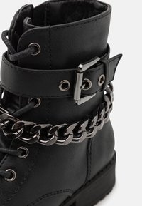 Černé faux kožené kotníkové boty s výrazným stříbrným řetízkem, detaily s přezkou a kulatou špičkou. Texturovaný svršek se šněrováním.