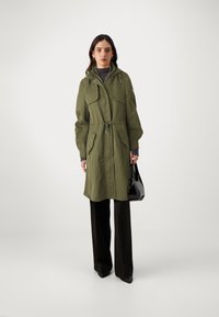 Cappotto lungo verde oliva con vita regolabile, cappuccio e due tasche frontali. Abbinato a pantaloni neri a gamba larga e una borsa nera.