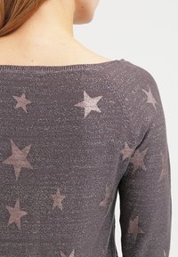 Grå stickad tröja med en avslappnad halsringning, som har ett subtilt stjärnmönster i ljusare nyanser och en texturerad yta.