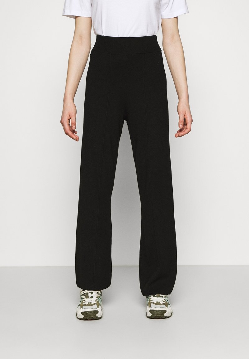 Personne portant un pantalon noir taille haute à jambes larges, une chemise blanche à manches courtes et des baskets blanches et vertes, debout devant un fond uni.