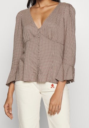 Kobieta ubrana w brązowo-białą koszulę w kratę z dekoltem w serek i rozszerzanymi rękawami oraz białe spodnie z małym czerwonym haftowanym detalem.