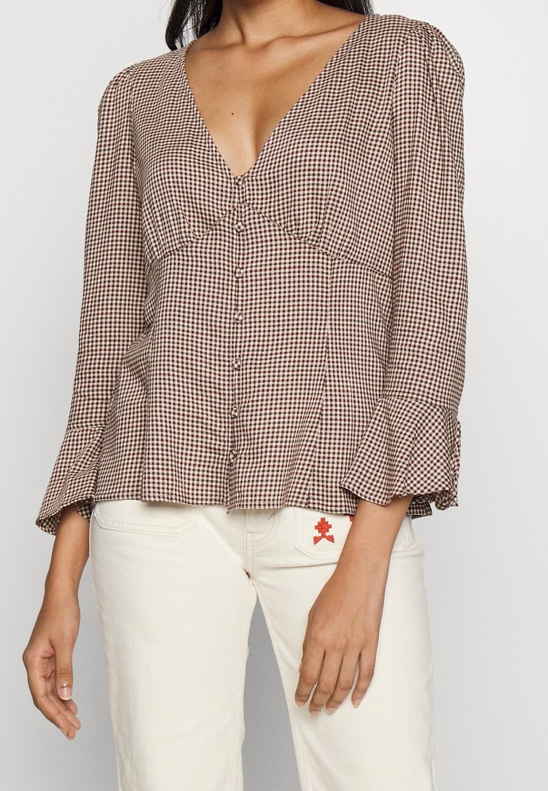 Femme portant un chemisier à col en V à carreaux marron et blanc avec des manches évasées et un pantalon blanc avec un petit détail brodé rouge.