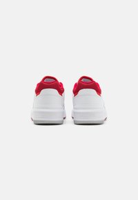 Valged sneakers punaste võrkkattega aktsentidega, millel on tekstureeritud tald ja "LACOSTE" bränding. Kujundus sisaldab ümardatud kontsa ja pehmendatud kaela.