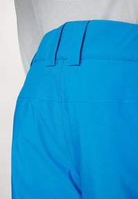 Blaue Hose mit glatter Textur, flachem Bund mit doppelten Schlaufen und kontrastierenden Nähten in der Nähe der Taschen.