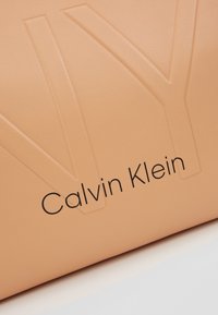 Pevný poutek s broskvovým texturováním. Embosovaný vzor "NY" a logo "Calvin Klein" v černé barvě, s hladkým povrchem z syntetické kůže.