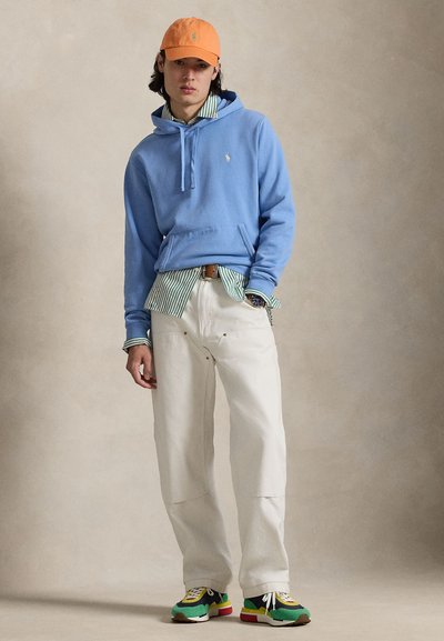 Γαλάζιο φούτερ με τσέπη καγκουρό, πράσινο και λευκό ριγέ πουκάμισο από κάτω, λευκό παντελόνι και πολύχρωμα αθλητικά παπούτσια. Πορτοκαλί καπέλο στο κεφάλι.