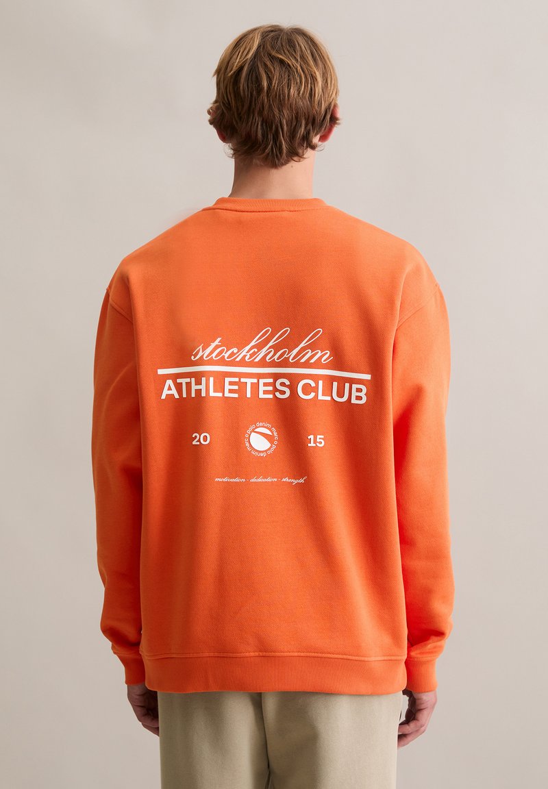 Pomarańczowa bluza z okrągłym dekoltem, z białym napisem "stockholm ATHLETES CLUB" i okrągłym logo; miękka w dotyku, o luźnym kroju.