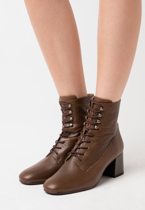 Bottines en cuir marron avec talons carrés et lacets portées pieds nus sur un fond blanc.