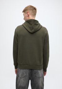 Bluza z kapturem w kolorze oliwkowej zieleni, z sznurkiem w kapturze, ściągaczami na mankietach i w pasie, zestawiona z szarymi dżinsami z tylnymi kieszeniami.