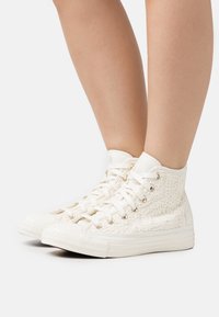 Nohy obuté do bílých vysokých tenisek s tkaničkami po vzoru krajky a bílými tkaničkami, zobrazené na jednobarevném bílém pozadí.