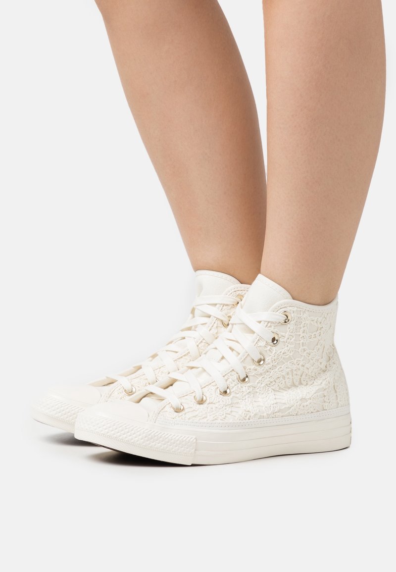 Nohy obuté do bílých vysokých tenisek s tkaničkami po vzoru krajky a bílými tkaničkami, zobrazené na jednobarevném bílém pozadí.